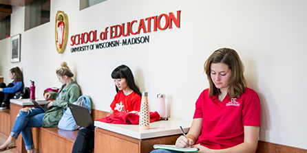 Students lined up by wall sitting down and working under School of Education sign.