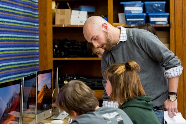 Teacher holding a paper, leans over to talk to students on MAC computers.