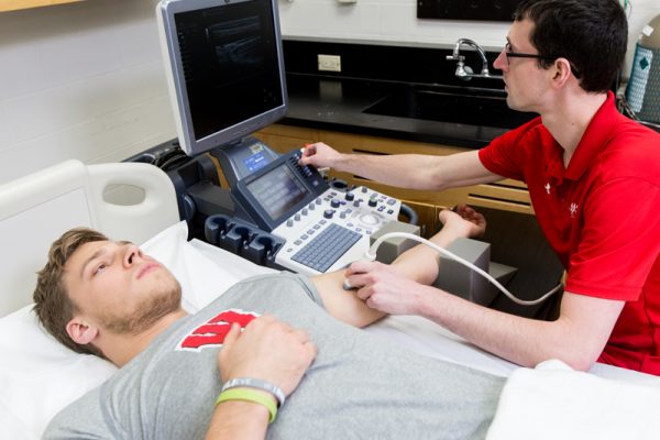 A student in a red shirt studies another student who is laying down on a bed for health research.