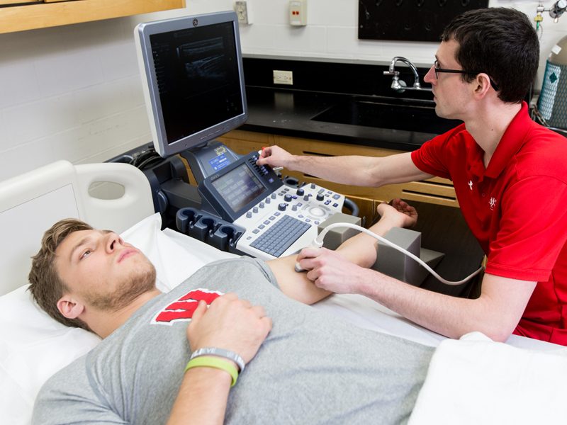 A student in a red shirt studies another student who is laying down on a bed for health research.