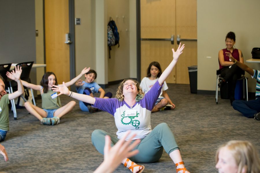 Teacher sitting on floor looking up with arms open surrounded by smiling students.