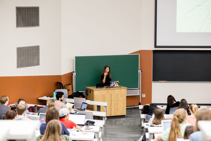 Professor standing in front of green chalkboard in large lecture hall.