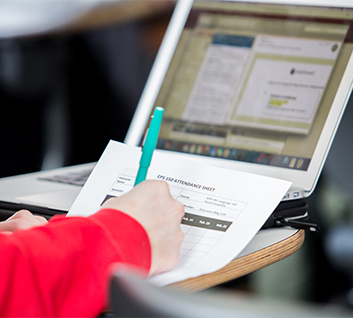 Student writing down on paper with blue pen and laptop on desk.