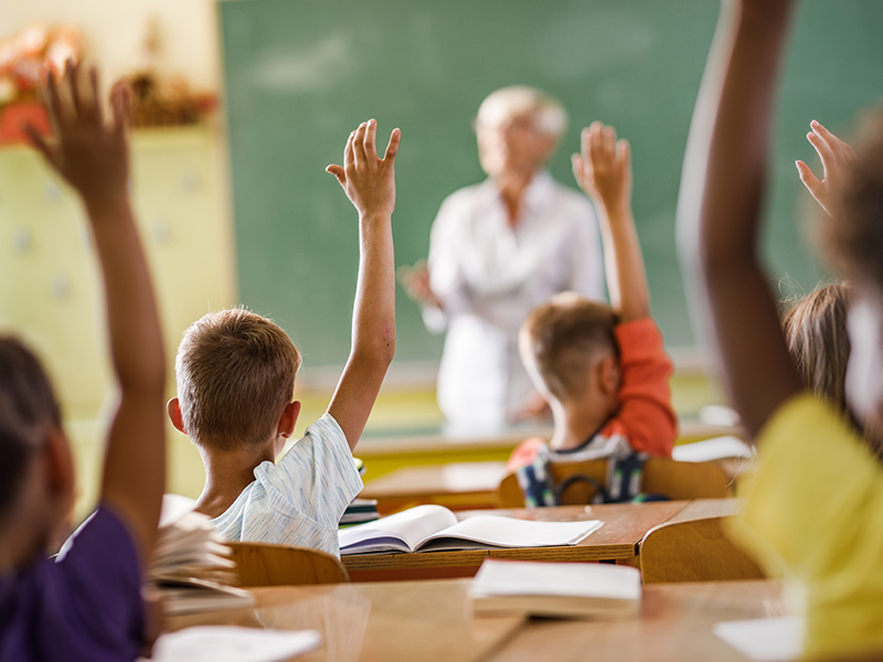 Young students raising their hands in class with their books on the desks.