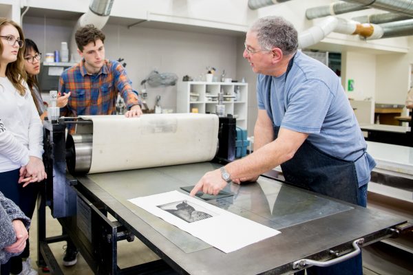 Professor leaning over printing table and demonstrating to students who are gathered around.